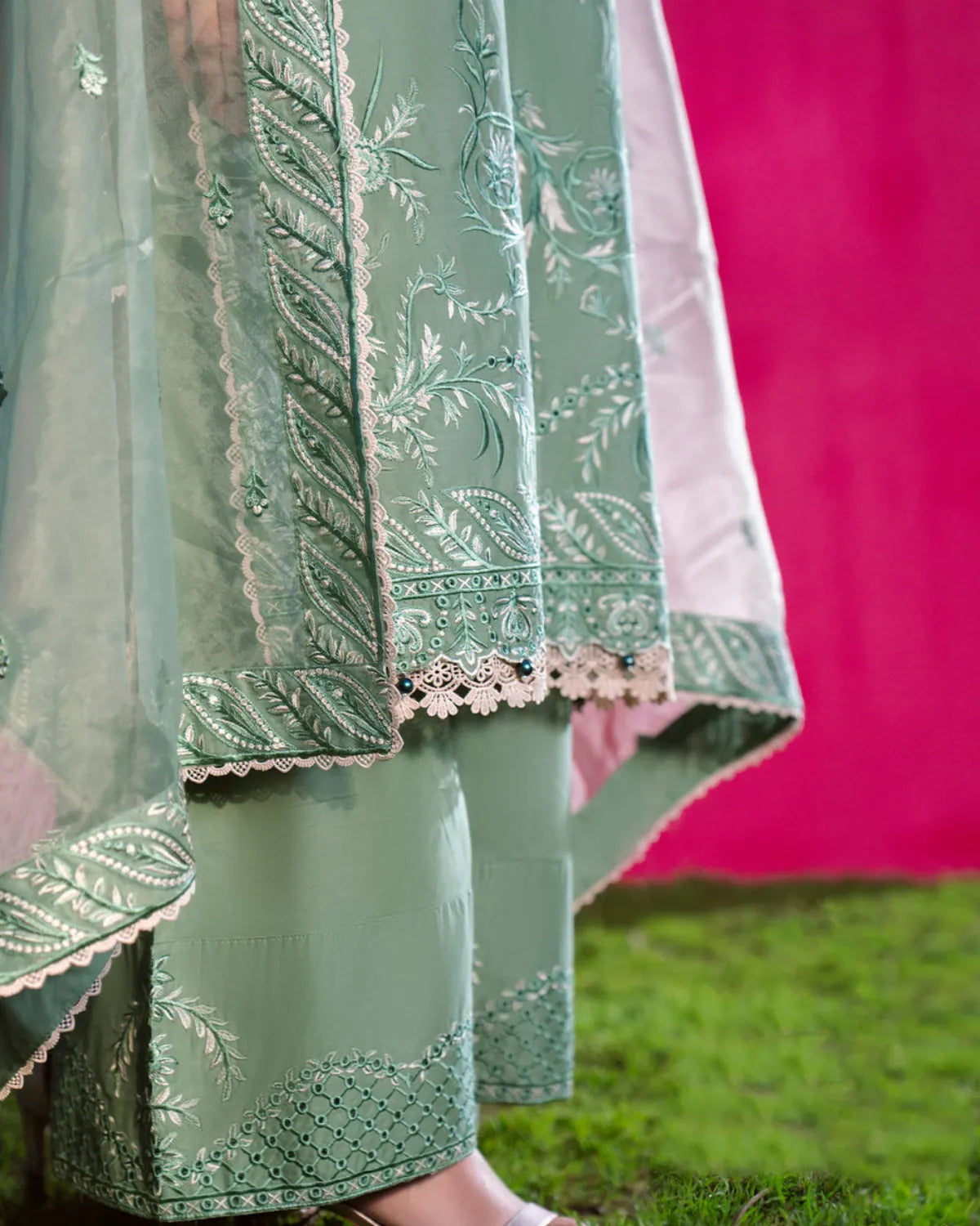 A close-up of a mint green embroidered traditional outfit with matching dupatta and wide-leg pants, featuring intricate floral embroidery and lace borders, shown outdoors against a bright pink wall and green grass.