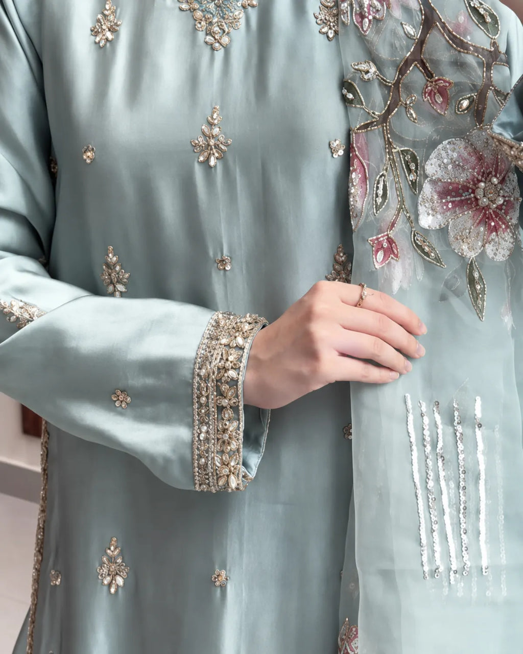 A close-up of a person wearing a light sage green traditional outfit with intricate gold and silver embroidery and a matching sheer dupatta featuring large pink and white floral motifs.