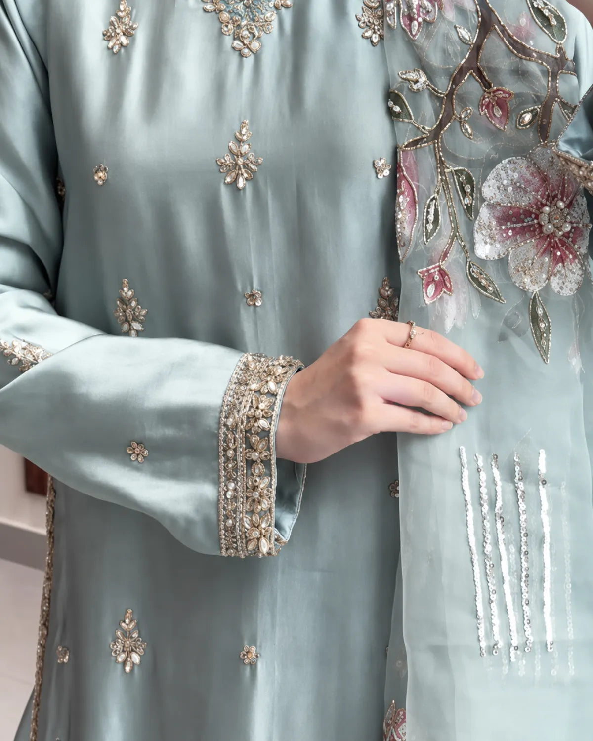 A close-up of a person wearing a light sage green traditional outfit with intricate gold and silver embroidery and a matching sheer dupatta featuring large pink and white floral motifs.