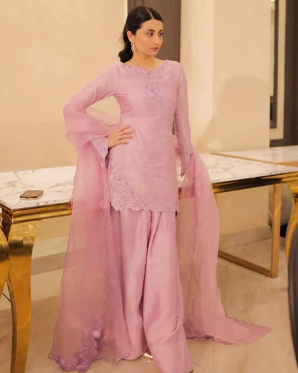 A woman modeling a light pink traditional South Asian outfit with a long kurta, wide flared sleeves, matching wide-leg pants, and a sheer dupatta, standing in an indoor setting with marble-topped tables.
