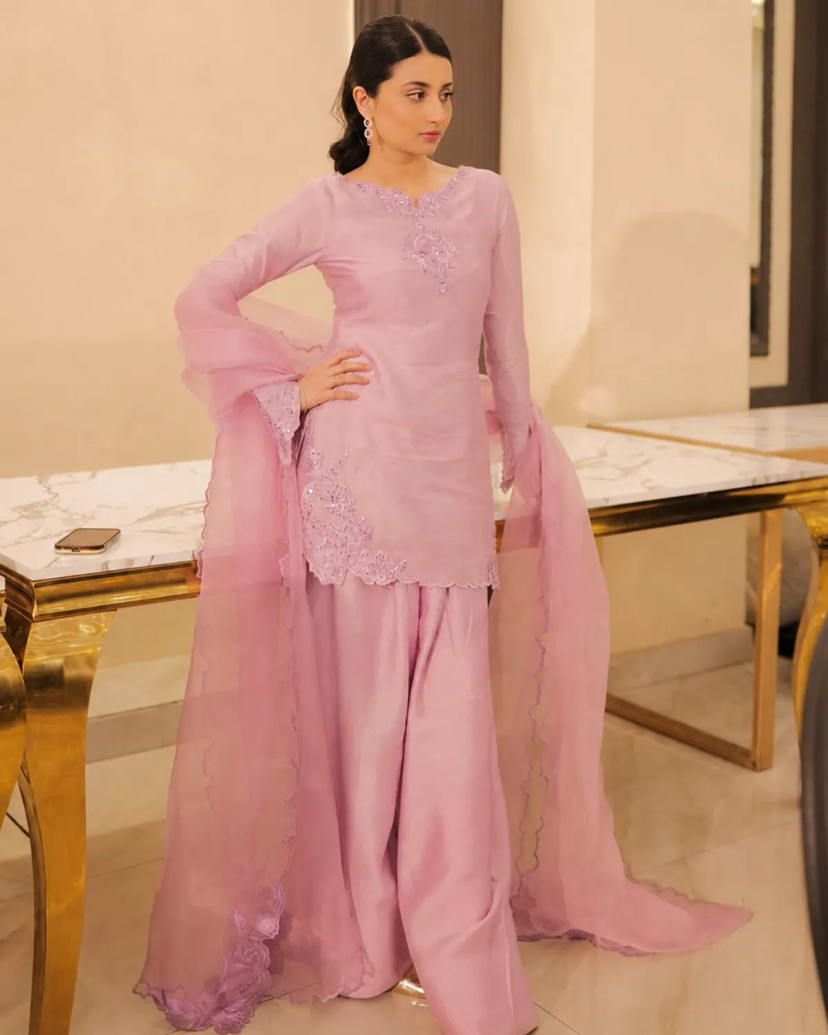 A woman modeling a light pink traditional South Asian outfit with a long kurta, wide flared sleeves, matching wide-leg pants, and a sheer dupatta, standing in an indoor setting with marble-topped tables.