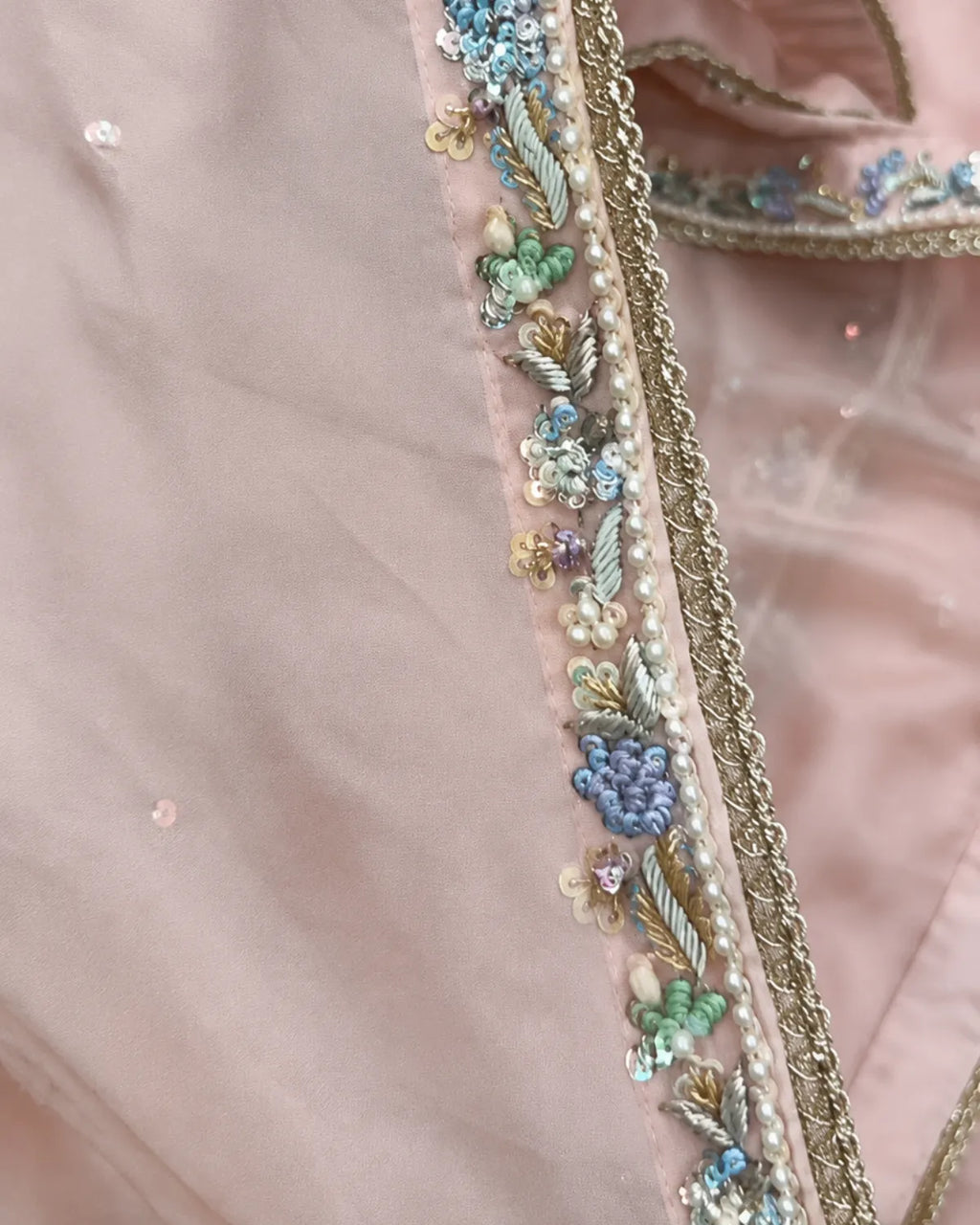 Close-up of a light pink fabric garment with ornate multicolor embroidery and beadwork along a border, including blue, green, gold, and pearl-like details.