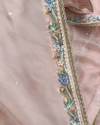 Close-up of a light pink fabric garment with ornate multicolor embroidery and beadwork along a border, including blue, green, gold, and pearl-like details.