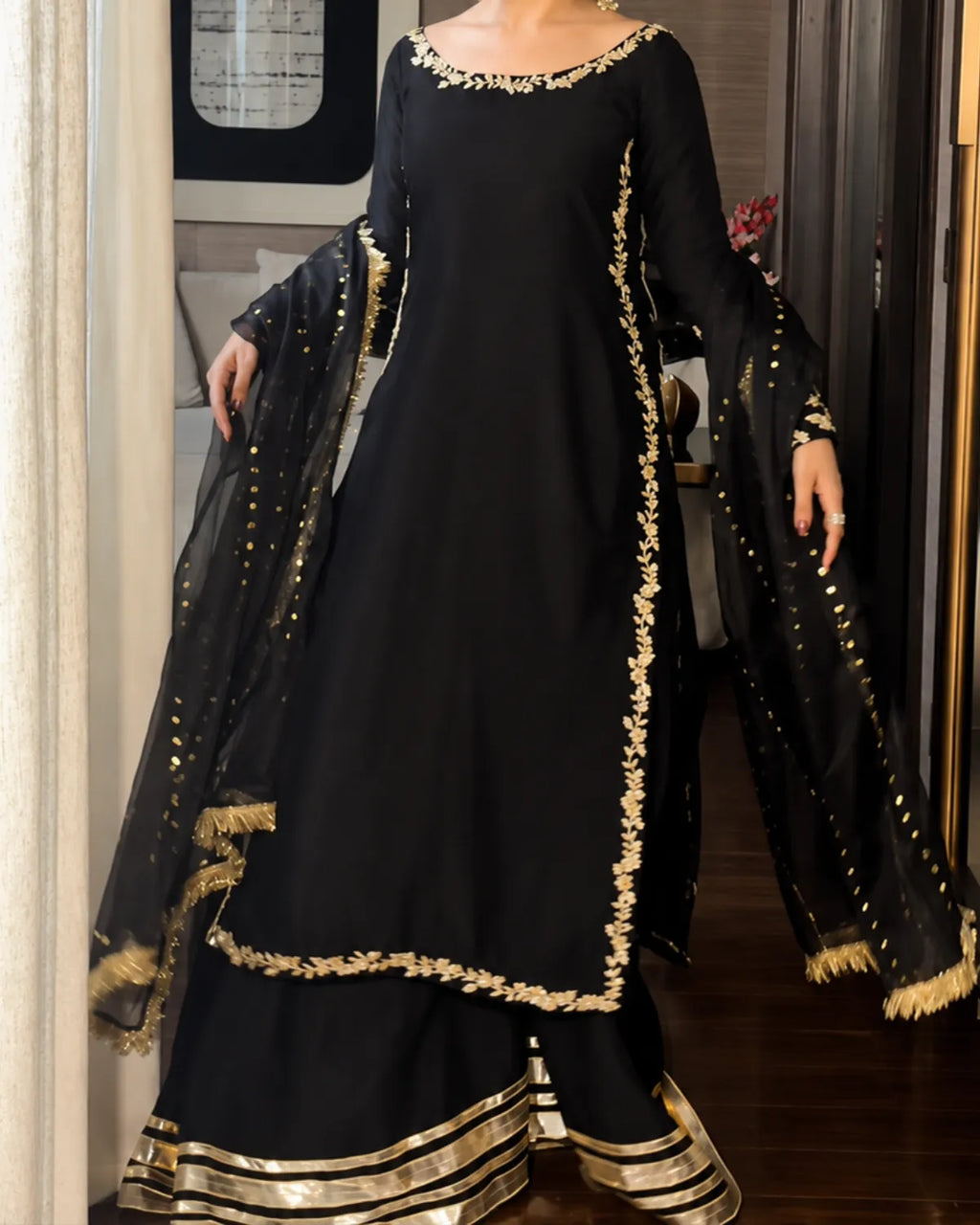 A woman modeling a black traditional South Asian outfit with gold embroidery, featuring a long kurta, flared pants, and a sheer dupatta draped over her arms in an indoor setting.