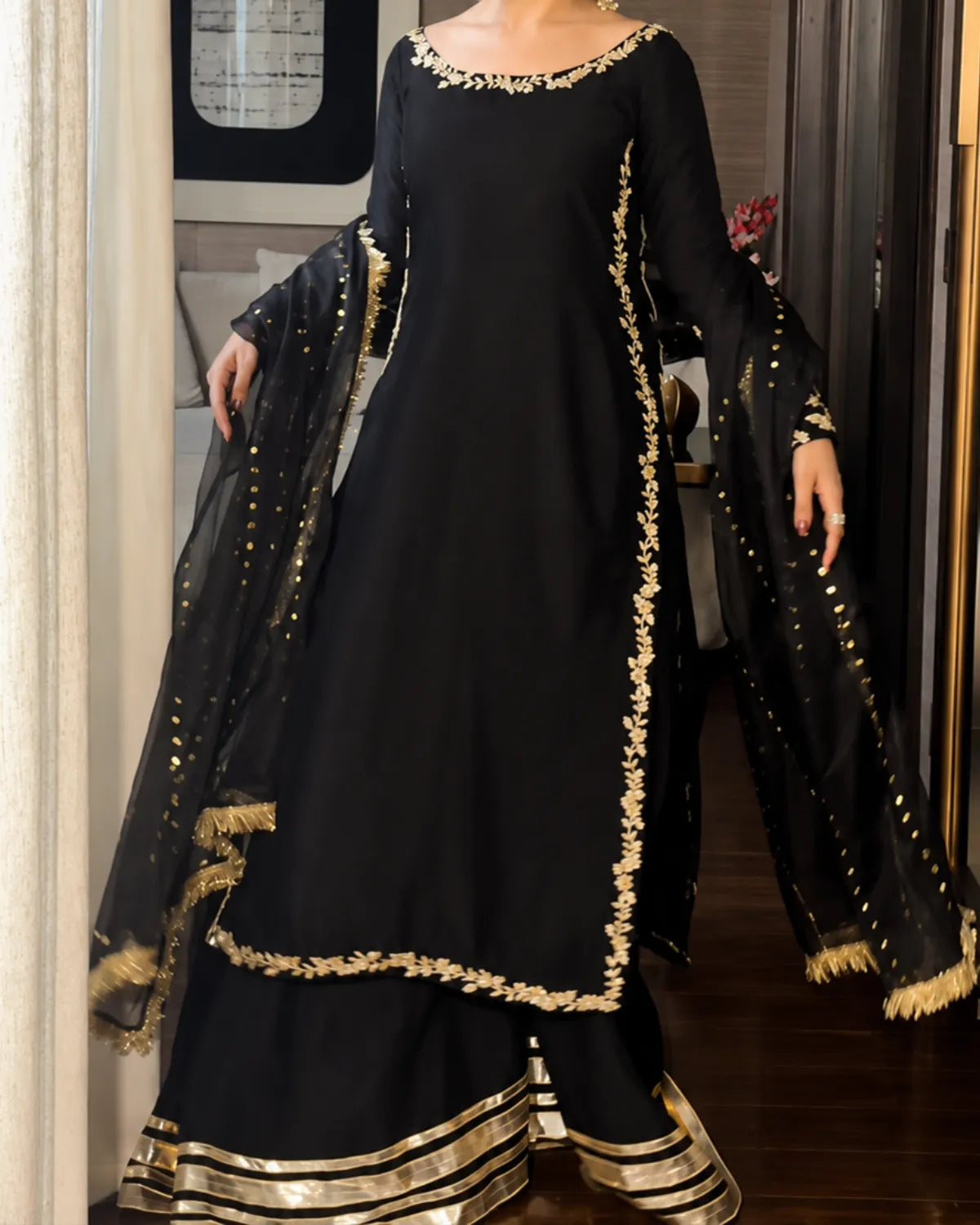 A woman modeling a black traditional South Asian outfit with gold embroidery, featuring a long kurta, flared pants, and a sheer dupatta draped over her arms in an indoor setting.