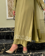 A person wearing a flowing olive green abaya or dress with embroidered lace trim at the hem and cuffs, paired with sparkly beige high heels, standing on black marble stairs indoors.