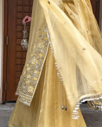 A close-up of a person wearing a flowing pale yellow traditional outfit with ornate silver and gold floral embroidery on the sleeve and border, paired with a sheer matching dupatta and holding a metallic clutch bag.