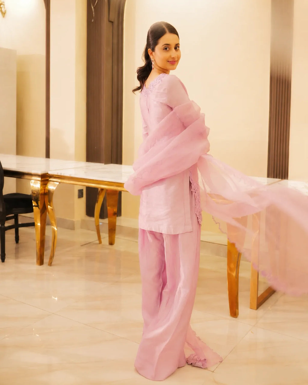 A woman wearing a light pink traditional outfit with matching sheer dupatta, standing in an indoor room with beige walls and a long table in the background.