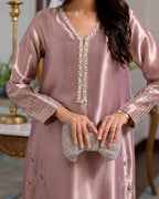 A woman wearing a satin-like dusty pink traditional tunic with ornate silver beaded trim along the V-neckline and cuffs, holding a matching beaded clutch in an indoor setting.