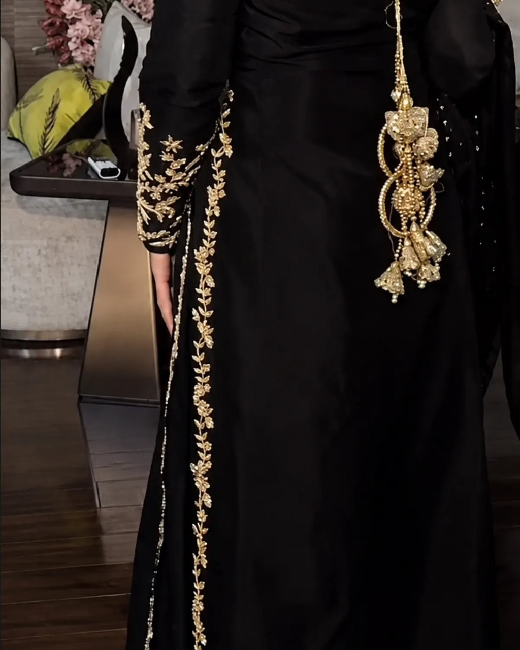 A close-up of a black traditional dress with ornate gold floral embroidery running down the side and sleeve, featuring a large hanging gold embellished accessory, shown in an indoor setting.