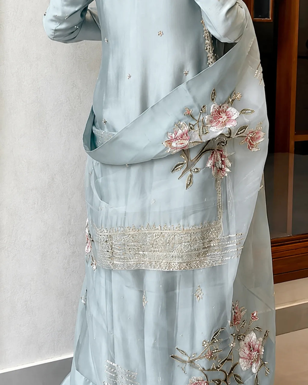 A light blue traditional outfit with pink floral embroidery and silver mirror work, shown on a model indoors.