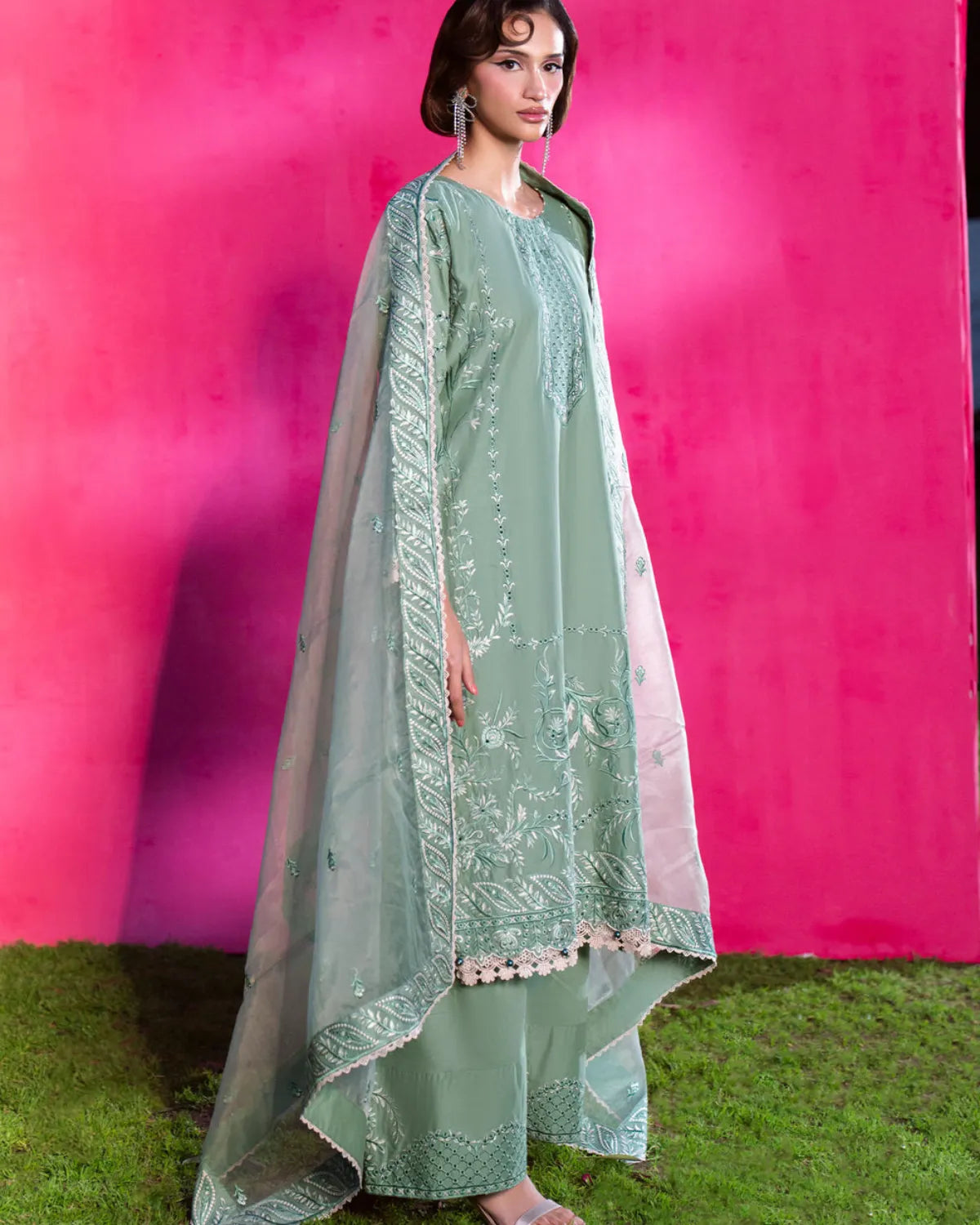 A woman models a traditional three-piece salwar kameez suit in sage green, featuring an embroidered kameez top, matching trousers, and a sheer dupatta with floral embroidery and pom-pom borders, set against a vibrant pink backdrop.