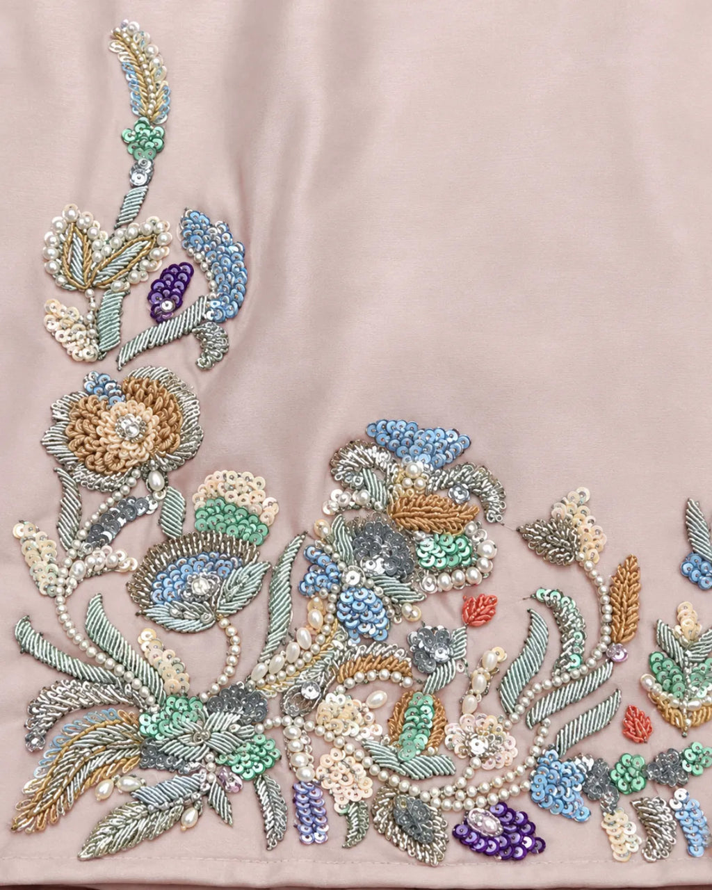 Close-up of a light blush pink fabric featuring intricate multicolor floral embroidery with sequins, beads, and pearl-like details concentrated along the bottom and left edges.