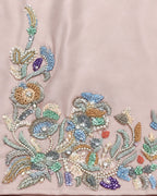 Close-up of a light blush pink fabric featuring intricate multicolor floral embroidery with sequins, beads, and pearl-like details concentrated along the bottom and left edges.