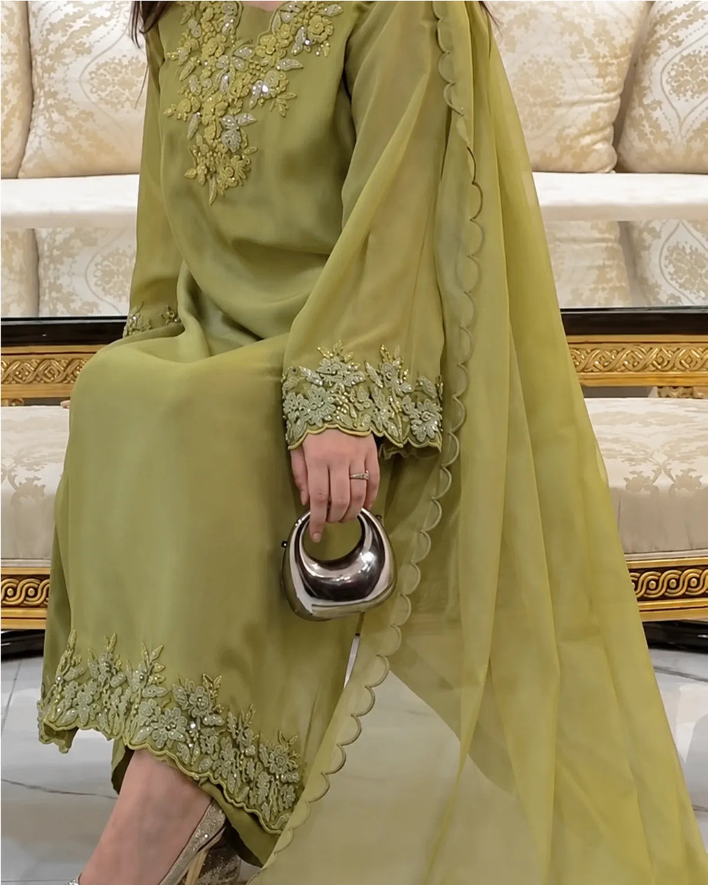 A woman wearing a sage green traditional outfit with floral embroidery and a matching sheer dupatta, holding a metallic silver crescent-shaped handbag in an indoor setting.