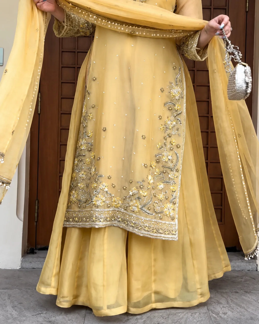 A person wearing a yellow traditional outfit with silver and gold embroidery, holding a matching sheer dupatta and a small silver handbag, standing in front of a brown wooden door.