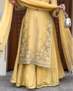 A person wearing a yellow traditional outfit with silver and gold embroidery, holding a matching sheer dupatta and a small silver handbag, standing in front of a brown wooden door.