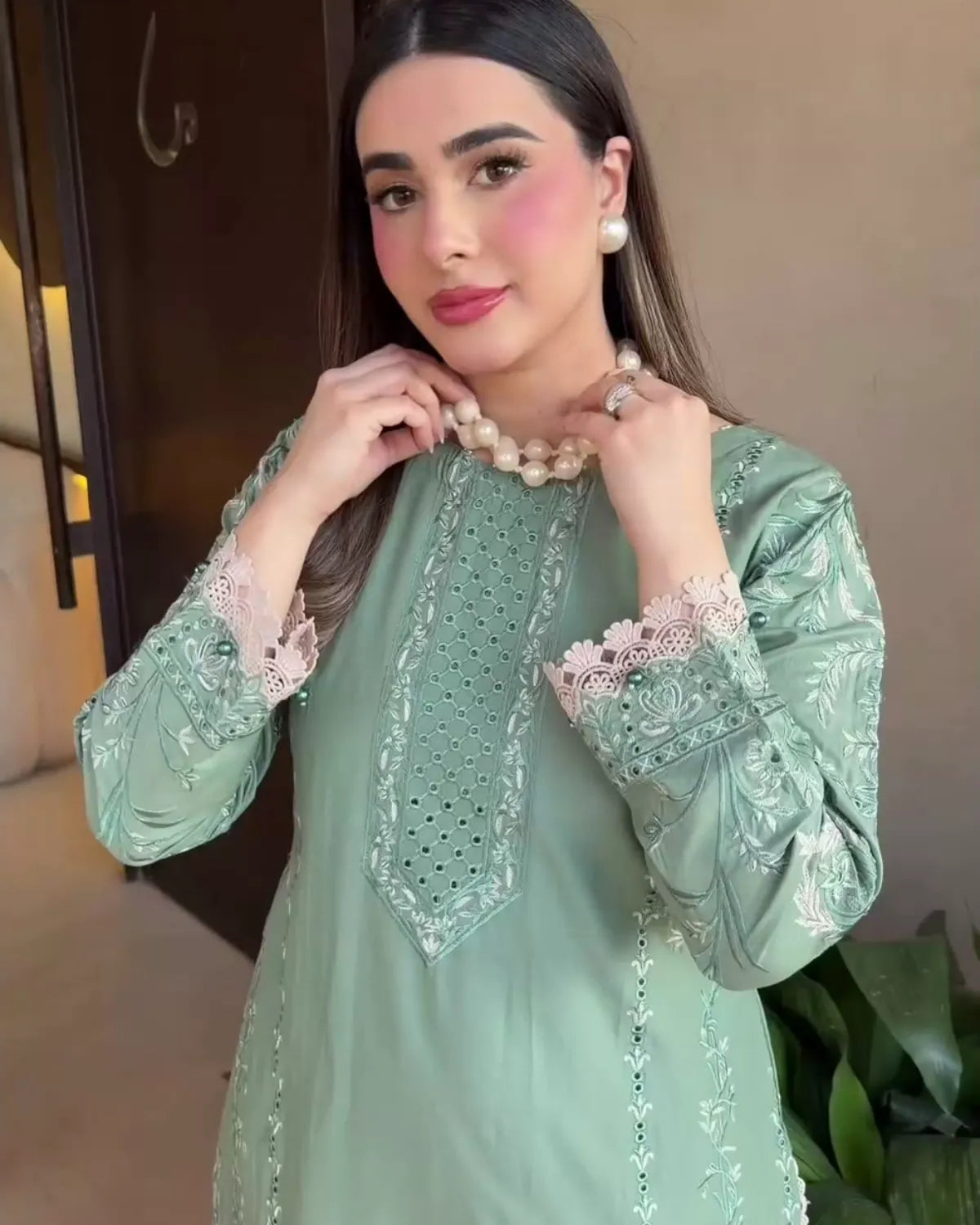 A woman wearing a sage green embroidered tunic with lace cuffs and a pearl necklace, posing indoors.