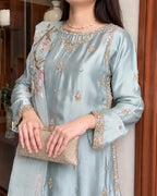 A woman wearing a light blue satin kurta with gold and silver embroidery, holding a metallic clutch purse in an indoor setting.