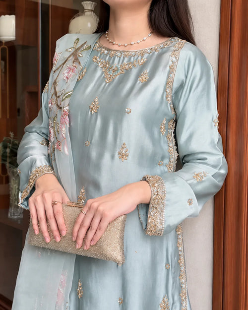 A woman wearing a light blue satin kurta with gold and silver embroidery, holding a metallic clutch purse in an indoor setting.