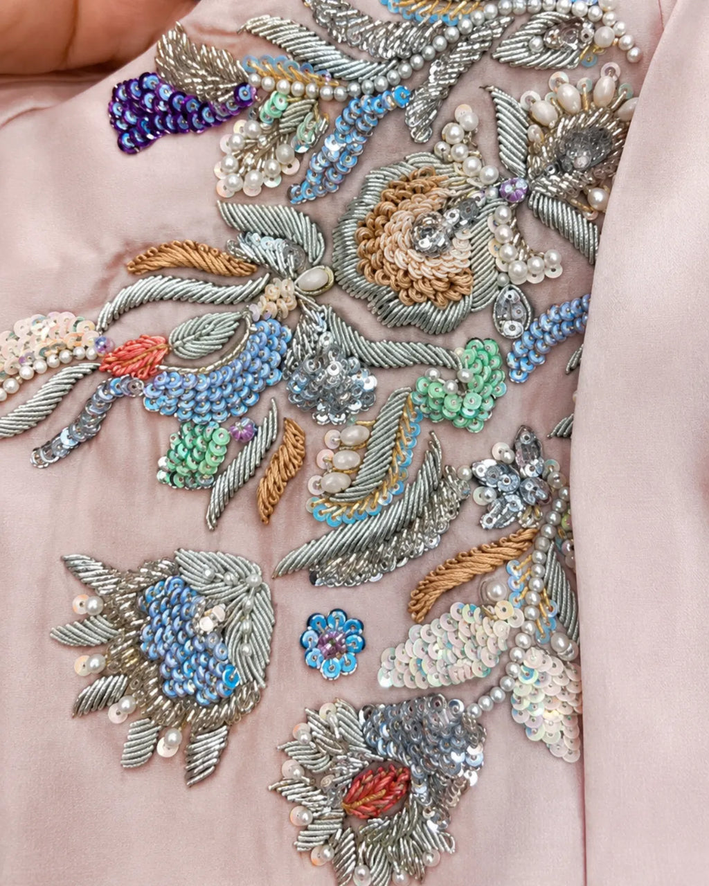 Close-up of a light pink fabric featuring elaborate multicolor embroidery with silver thread, sequins, beads, and pearl-like details.