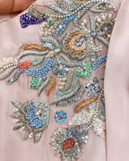 Close-up of a light pink fabric featuring elaborate multicolor embroidery with silver thread, sequins, beads, and pearl-like details.