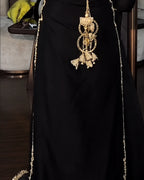 A close-up of a black dress or abaya with ornate gold embroidery and a large dangling gold embellishment at the center, shown against an indoor background.