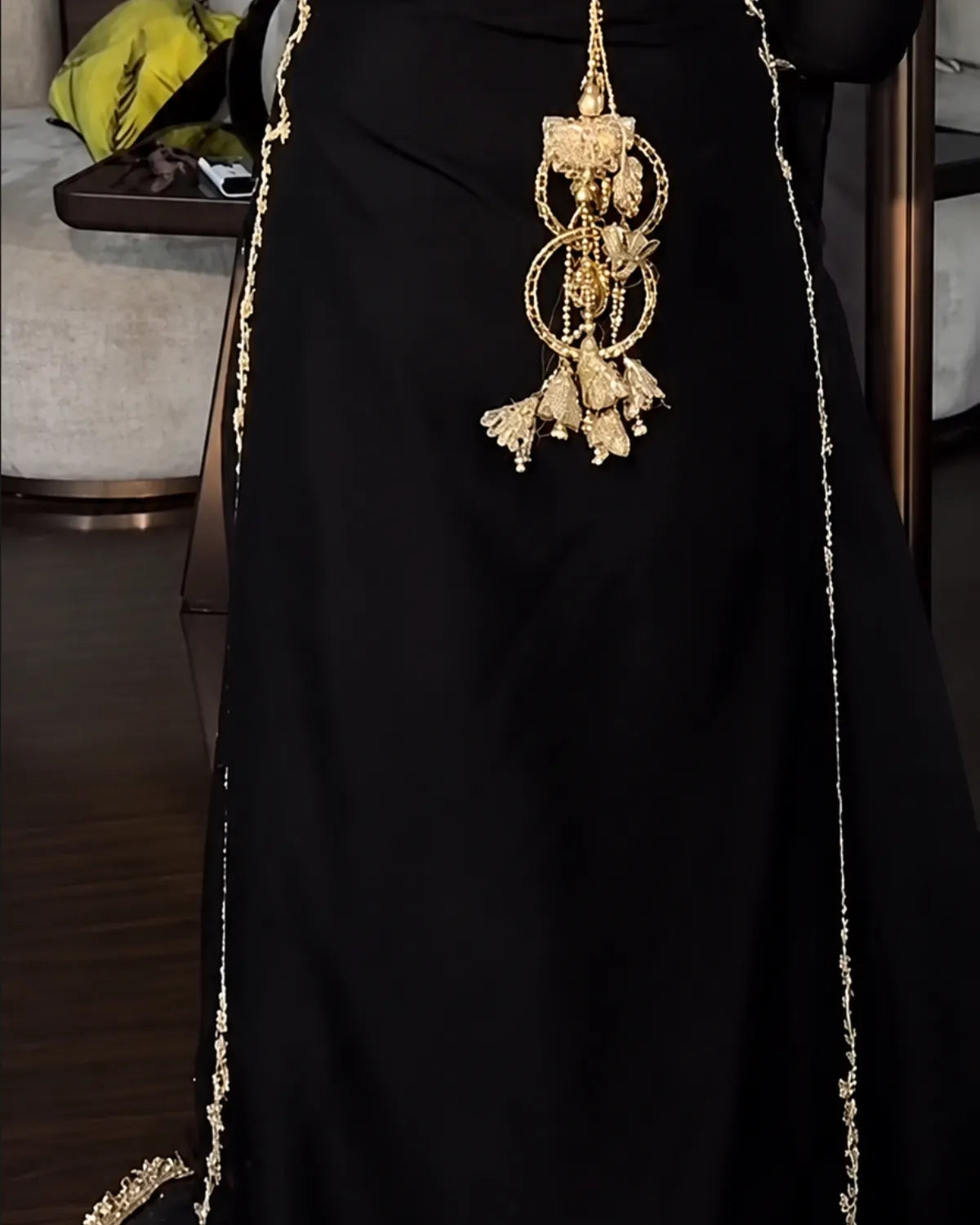 A close-up of a black dress or abaya with ornate gold embroidery and a large dangling gold embellishment at the center, shown against an indoor background.