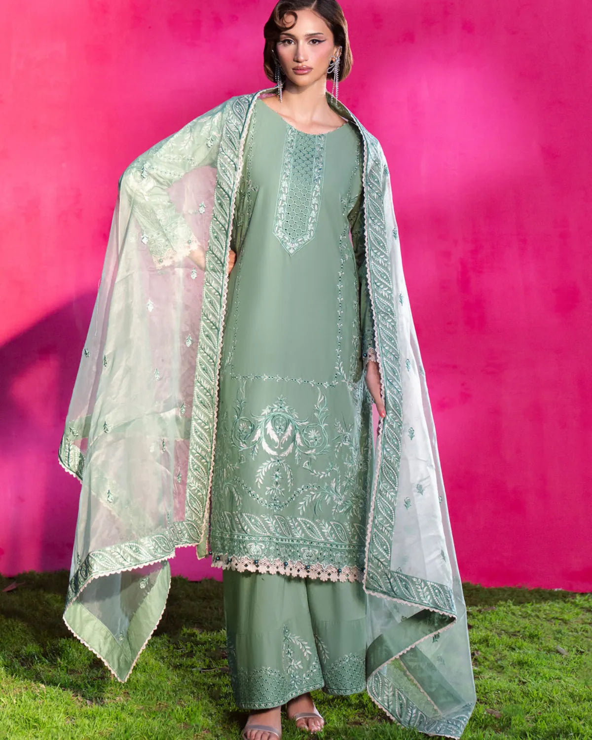 A woman models a sage green embroidered traditional outfit with a matching sheer dupatta, set against a bright pink wall and green grass.