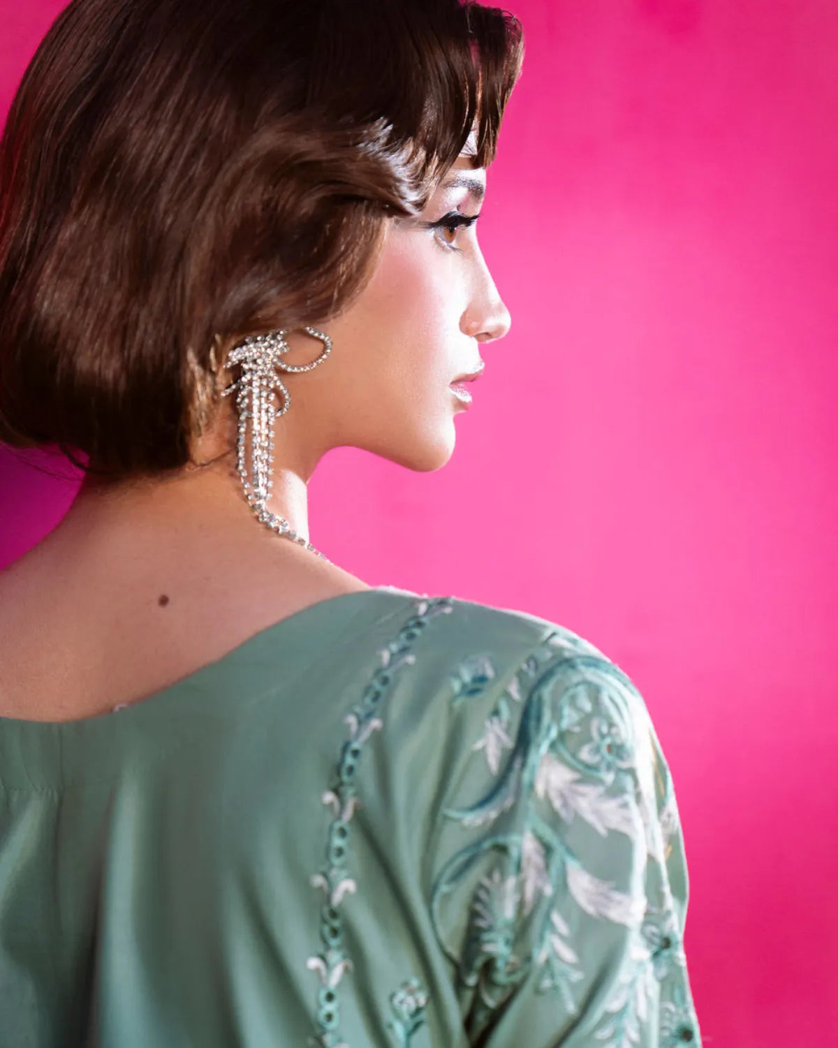 A woman in profile wearing a sage green embroidered blouse and a long dangling silver beaded earring, set against a vibrant hot pink background.
