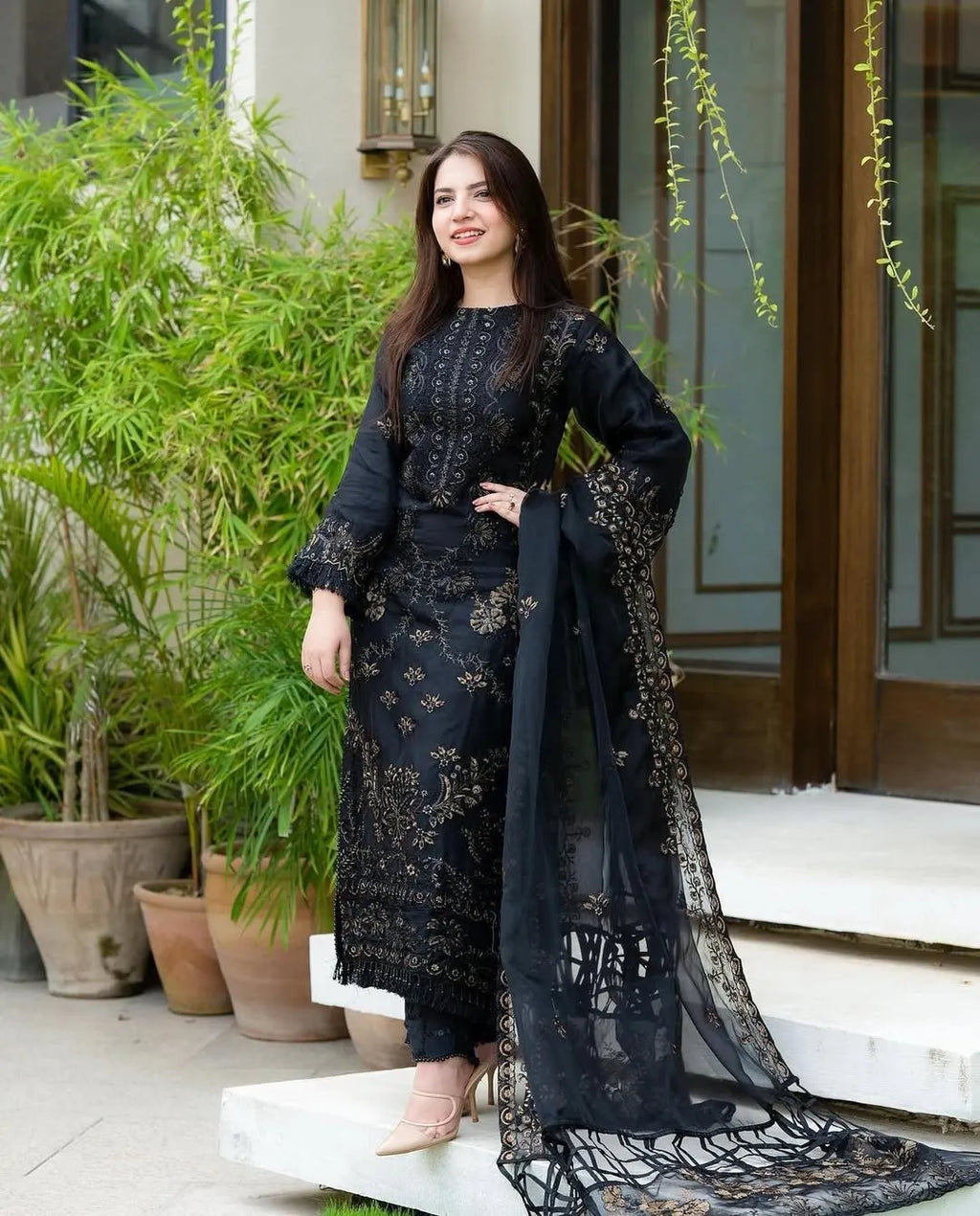 Woman in a black embroidered outfit standing outdoors with plants and a door in the background