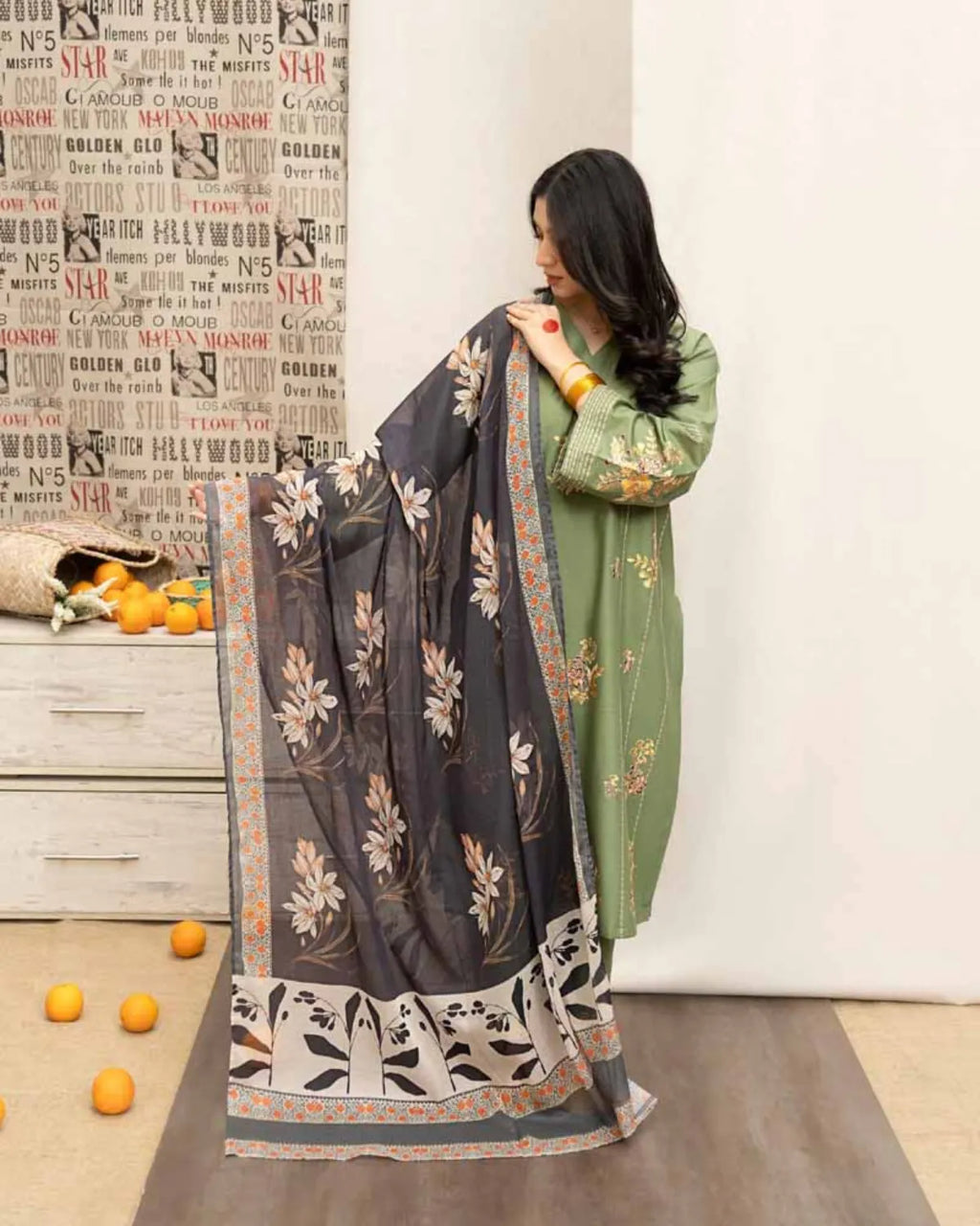 Woman holding a floral-patterned saree in a room with a newspaper wall and oranges on the floor.