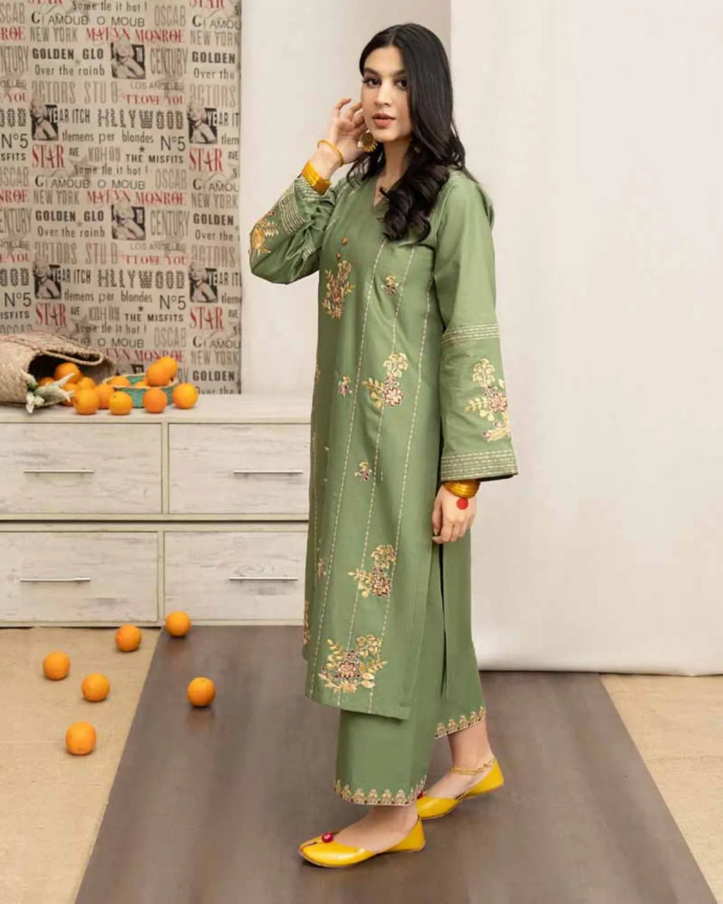 Woman in a green embroidered outfit standing in a room with a decorative wall and scattered oranges on the floor.