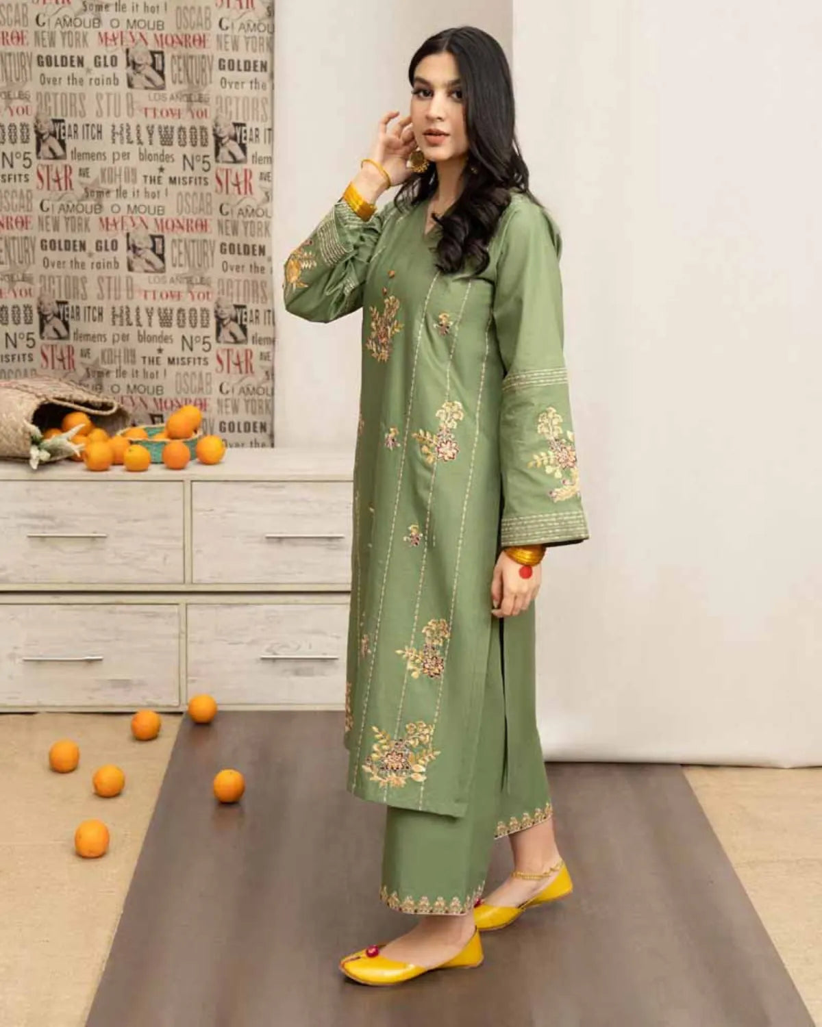 Woman in a green embroidered outfit standing in a room with a decorative wall and scattered oranges on the floor.