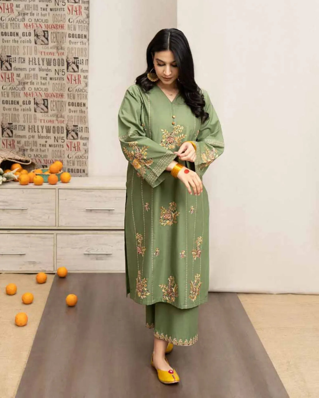 Woman in a green embroidered outfit standing in a room with a newspaper wall and scattered oranges on the floor.