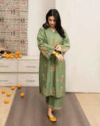 Woman in a green embroidered outfit standing in a room with a newspaper wall and scattered oranges on the floor.