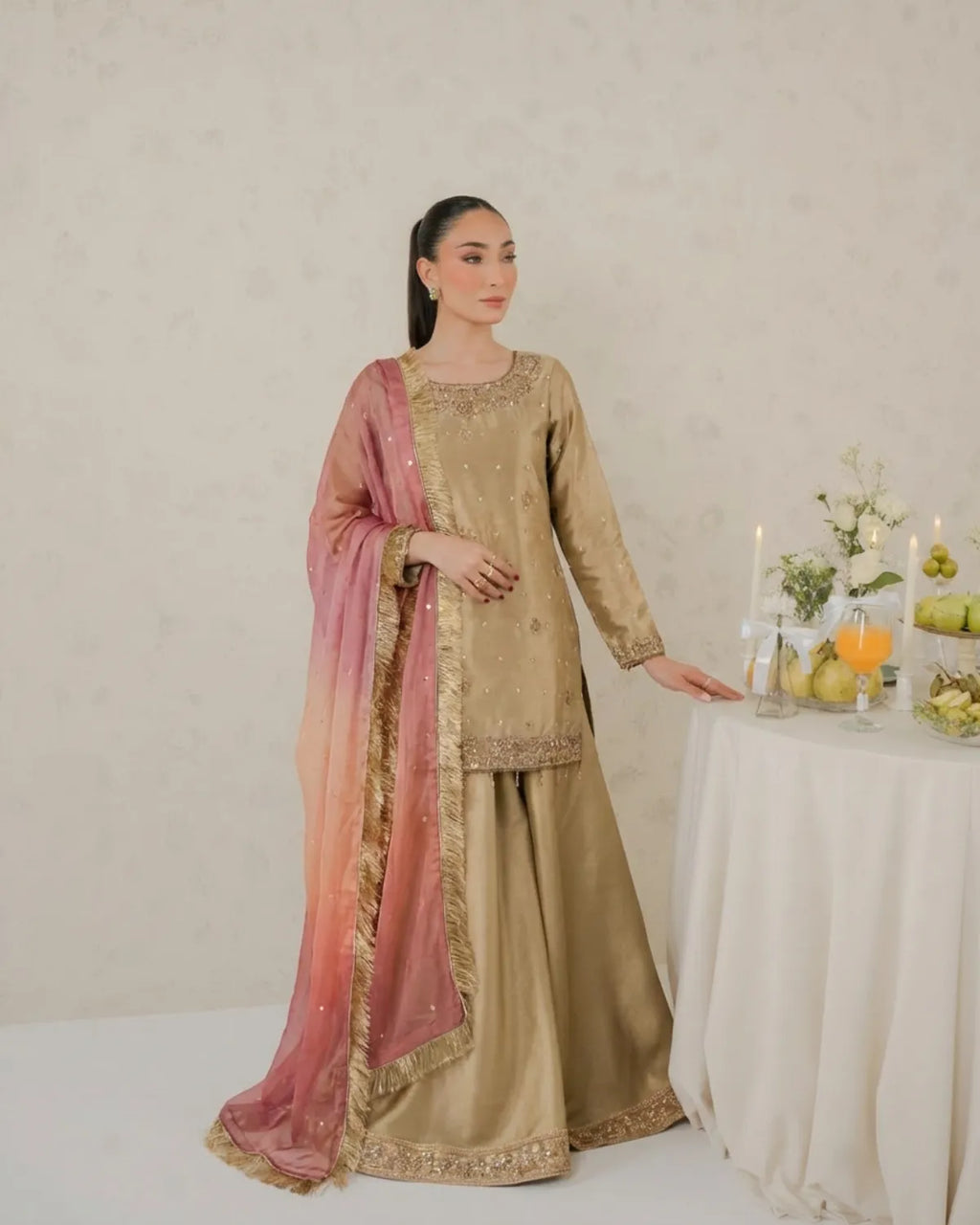 Woman in a traditional outfit with a pink dupatta standing next to a table with fruits and candles.