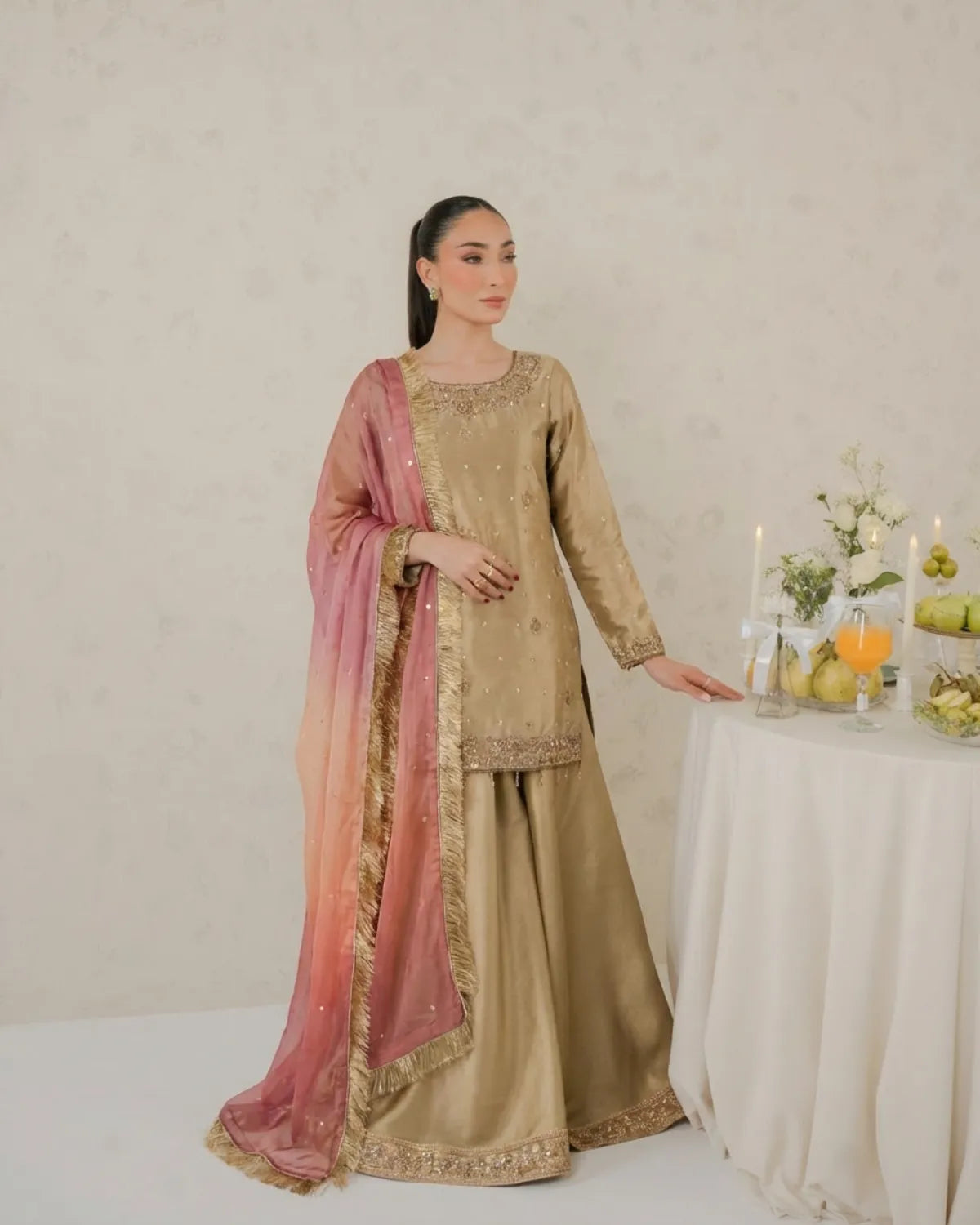 Woman in a traditional outfit with a pink dupatta standing next to a table with fruits and candles.