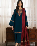 Woman wearing a teal and red traditional outfit with embroidery, standing in front of a wooden cabinet.