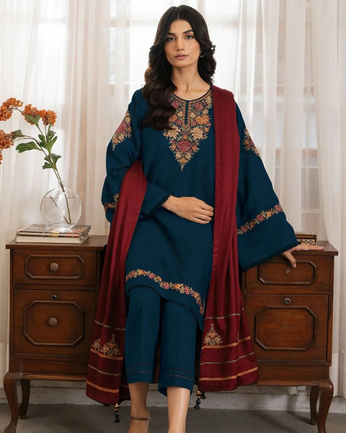 Woman wearing a blue embroidered outfit with a red dupatta in a room with white curtains and a wooden side table.