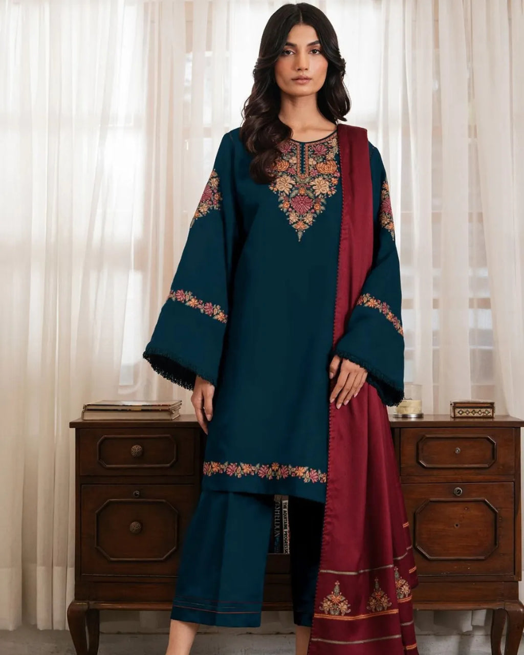Woman wearing a teal and red traditional outfit with embroidery, standing in front of a wooden cabinet.