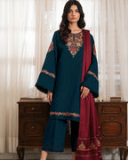 Woman wearing a teal and red embroidered traditional outfit with a wooden cabinet in the background.