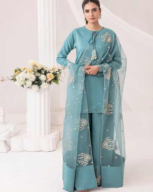 Woman wearing a teal traditional outfit with floral patterns in a white room.