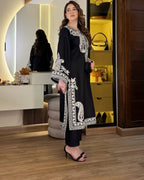 Woman in a black and white embroidered outfit standing in a room with a mirror and shelves.