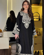 Woman wearing a black dress with white embroidery in a room setting.