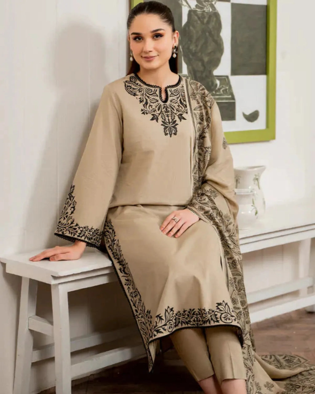 Woman wearing a beige embroidered outfit sitting on a stool indoors.