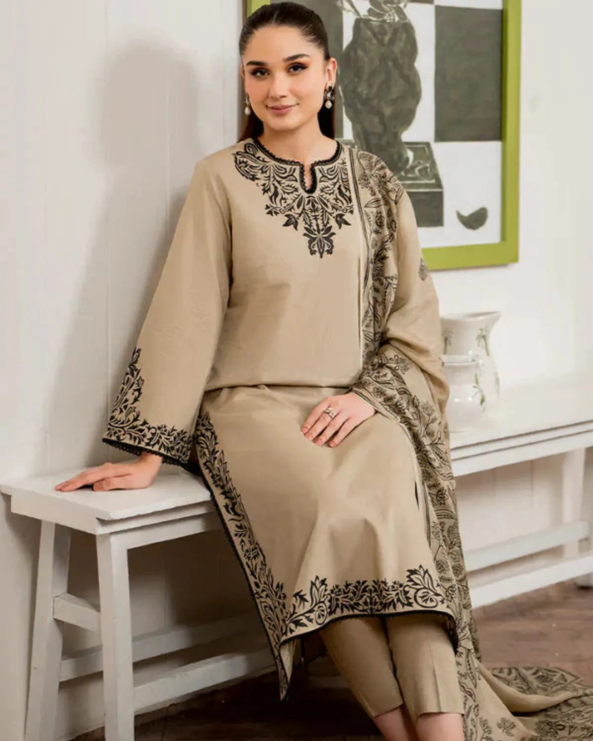 Woman wearing a beige embroidered outfit sitting on a stool indoors.