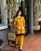 Woman in a yellow outfit standing in an indoor setting with plants and wooden furniture.